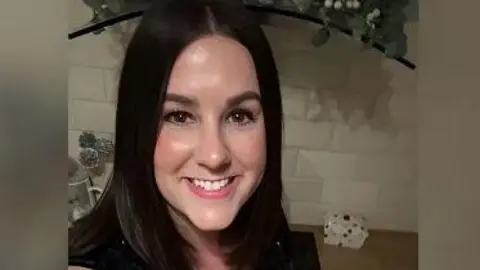 Sourced from X Lucy Connolly, who has dark, shoulder length hair is smiling at the camera. She is standing in front of a white tiled wall indoors.