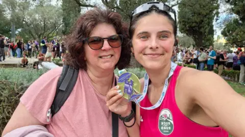 Claudia Andrade Claudia and Idalina are standing close together outdoors at a large public event. Claudia holds a round medal on a ribbon toward the camera. A crowd of people and tall trees fill the background.