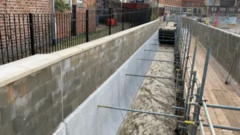 The Environment Agency The brook runs through the city and a raised wooden ledge runs along the right side of it with scaffolding holding it up. Metal poles are touching the opposite side of the brook where new higher walls have been put in place. The wall is part smooth concrete then rows of bricks have been added. Housing can be seen on the left.