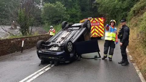 Wide shot of an overturned car. There is a police officer and another emergency worker beside the vehicle. The road is obviously wet.