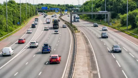 Cars travel north and south on a motorway with a junction exit on the left of the image. Overhead signs can be seen in the distance.