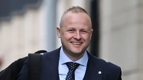 Jamie Bryson is wearing a dark suit and tie and is smiling as he walks to a court hearing. He has thinning fair hair and some stubble.