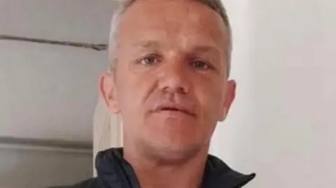 Peter Phillips has close cropped grey hair and is wearing a black sleeveless jacket with a white t-shirt underneath. He is standing in a domestic kitchen.