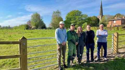 BBC Four men and one woman stand by a metal gate attached to wooden fencing. There is a large grassy field behind them backing onto a graveyard. Trees, a house and a church can be seen in the distance.