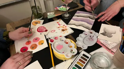 Holly Nichols/BBC A small table is covered with art supplies and watercolour paintings in progress. Several hands are visible as people paint colourful fruit designs, a pink pomegranate, soft purple landscape-style waves, and a small grey figure. Paint palettes, brushes, paper towels with test marks, and drinks are scattered around the table.