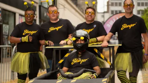 Ice Eye Media Four team members and their driver stand next to their kart wearing yellow and black T'shirts, socks and netting as skirts
