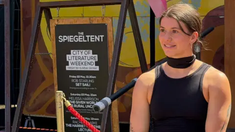 James McConnell/BBC Christina stands in front of a sign advertising other acts on at the festival over the weekend. She is smiling, looking out of frame, and wears a black vest and fashion scarf. Her hair is brown and pulled back.