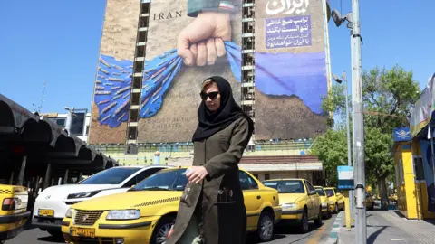 A woman walks through Tehran's Vanak Square with a poster referring to the Strait of Hormuz in the background and yellow taxis on the street