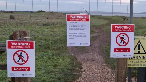 New Forest District Council A wire fence across the gateway to a gravel path which meanders into the distance along a grassy clifftop. The sea and coast are in the distance. Three signs are attached to the fence saying, warning footpath closed and no entry.