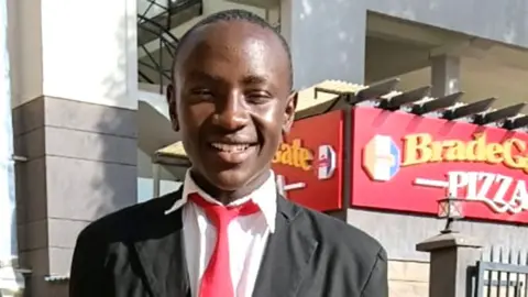 A photograph of Sylvester Muigai Ndung'u in a school uniform. He is in a grey jacket, white shirt and red tie.