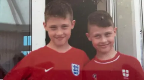 Family Two twin boys with short brown hair and wearing red England football shirts smiles as they pose for a photo outside a door.