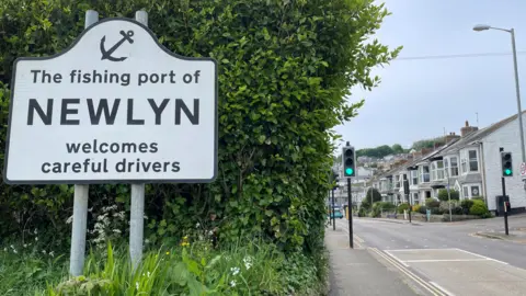 BBC The welcome sign of Newlyn. It says the fishing port of Newlyn welcomes careful drivers. A green bush is behind the sign. To the right of the sign is a view down a street in the town. A row of houses and traffic lights are visible. 