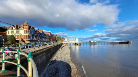 Getty Images Penarth sea front. The pier is seen at the back of the image, there is the sea in the foreground and to the right. On the left is the sea wall and there are Victorian buildings, but before those there is a road with cars parked on them.