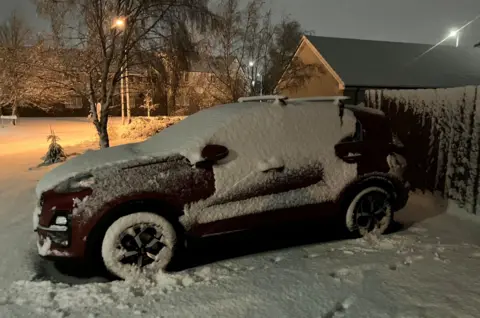 Snow covers a car. There is thick snow around it. It is early morning and streetlights are on.