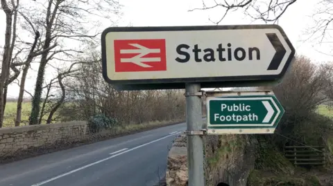 A road sign for a train station above a green public footpath sign on a pole by a roadside in Devon. It is next to a stone bridge in the countryside.