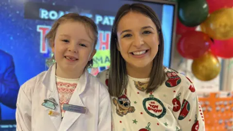 Roan St. Patrick's PS Zara is dressed in a white scientist coat, and standing beside her teacher who is wearing pyjamas