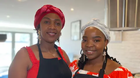 Tendai is to the left and Nyamwererei are in aprons standing side by side in a bright kitchen, facing the camera. Both are positioned in front of a cooker hood and tiled backsplash, with soft indoor lighting and kitchen items visible in the background