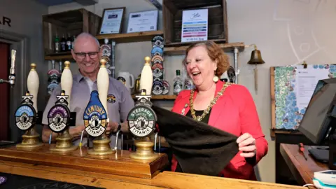 Lord Mayor Lyn Sharpe, wearing a red jacket and gold chains of office, is stood behind a bar with four pumps. She is stood next to a man with a grey shirt on.