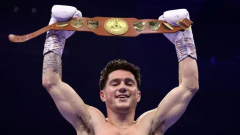 Mr Cooney with short brown hair wears white boxing gloves as he raises a brown boxing belt above his head.