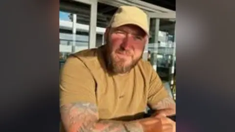 A man wearing a tan coloured baseball cap and t-shirt is sat looking into the camera. He has tattoos on both arms and a brown beard.
