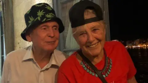 Family photo Professor Adrian Roscoe on the left is wearing a white shirt and a dark bucket sun hat with green leaves on. He’s looking at his wife Janice who’s blonde and wearing a baseball hat backwards and a red T Shirt. It was taken on holiday in Malta at night time. Both are smiling.