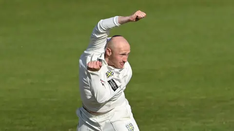 Left-arm spin bowler Callum Parkinson just after completing a delivery with his left arm tucked in and his right arm raised over his head 