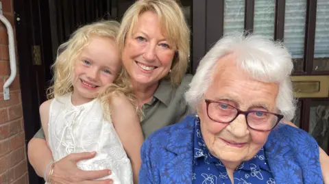 A young girl is in the arms of a woman, standing behind an elderly lady dressed in blue. They are all smiling at the camera.