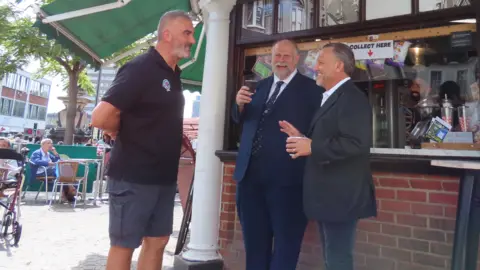 Bedford Borough Council Libby Lionetti, standing in front of his cafe, talking to Mayor Tom Wootton, holding a coffee cup and John McReynolds