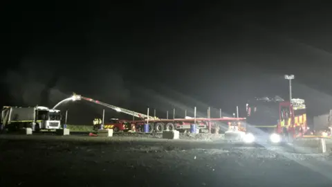 Brian Higginson The long crane from a fire service appliance is hosing water over a fire. There is some smoke in the background and a fire engine in the foreground with its lights on as it is dark.