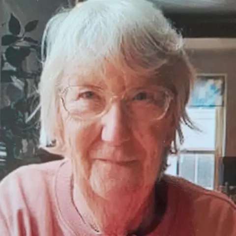 Helen Wilson An older woman with short white hair and clear rimmed glasses looks into the camera . She wears a pale pink jumper with the words 'be yourself' on. 