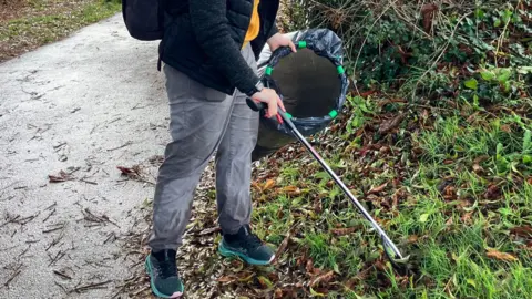 Generic image showing a woman litter-picking at a park. She holds a bin bag as she picks up rubbish with a litter-picking stick from a grassy area next to a path.