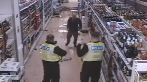 A CCTV still image showing a man in dark clothing walking down the Tesco aisle towards the camera, with contents of shelves and liquid spilt behind him. In the foreground, two male police officers have their back to the camera, facing the man.