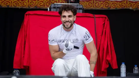 Getty Images Saint Levant is wearing all white a t-shirt and trousers, he is sat down on the stage holding a mic. He has dark curly hair and brown eyes, behind his is a red tablecloth. 