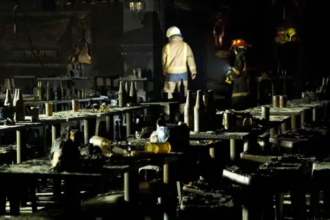 Getty Images Search and rescue teams inspect the interior of the bar, which is almost entirely charred. Blackened cups, bottles of beer and and beer buckets are seen on the tables
