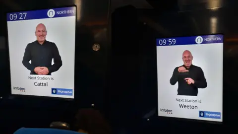 Northern Two screens on a Northern train show a sign language interpreter, wearing a black shirt, announcing the train's next stop.
