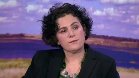 Claire Hanna - a woman with short, dark hair wears a dark suit jacket, a green shirt and a necklace. In the background is a screen displaying grass, water and a blue sky.
