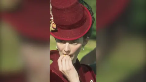 A woman with a maroon top hat and matching jacket puts a primrose in her mouth. She is standing in a field.