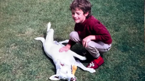 A grainy film photograph taken in the 1980s of a young boy playing on the grass with a white dog, who has a rubber bone in its mouth. The boy has short curly dirty blond hair and is wearing a red jumper, red slippers, and grey jogging bottoms. His eyes are closed and he is smiling.