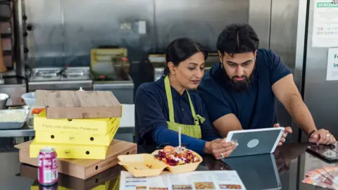 Getty Images Stock photo shows two business owners looking at a tablet at the counter of a takeaway restaurant with food boxes on the counter and cooking appliances in the background.