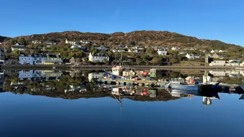 Allan McKellar A harbour on a bright winter day. The sky above is blue. The water below is calm and reflects the boats on the surface. A large hill is in the background with a row of colourful houses in blue, green, yellow and white in front of it. 