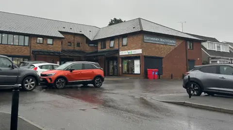 BBC News Llywynhendy health centre is a brick building with a sign. There is a car park in the foreground with several cars parked. 