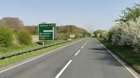 A dual carriageway with a sign on the left that says the A303 towards Andover. One car can be seen in the distance on the right and there are trees lining the verges.