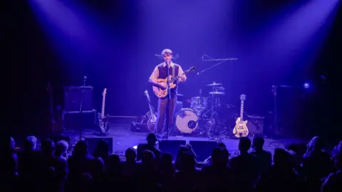 Plaster Communications Solo male musician on-stage singing and playing an electric guitar, with blue backlighting