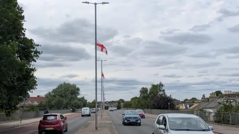 Alex Harris/BBC A row of lamp-posts in the middle of a dual carriageway. They all have St George's flags flying from them. Cars are driving on both sides of the road.