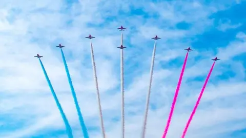 Several planes fly high in formation in the sky. They are spraying blue, white and pink smoke. 