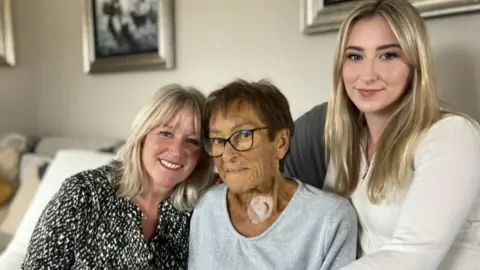 Shelley Crouch, her mother Maureen and daughter Mia are pictured sitting on a sofa. Shelly Crouch is wearing a black and white blouse and has blonde hair in a long bob. Maureen is wearing a grey, round-neck top and has a large circular plaster on her neck. Her hair is short and dark. Mia is wearing a cream top and has long blonde hair. All three are smiling.