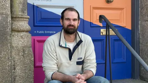 Felix has dark hair and a beard and wears a cream coloured jumper and jeans - he is sitting on some stone steps with brightly coloured painted doors heading into The Ladder behind him. 