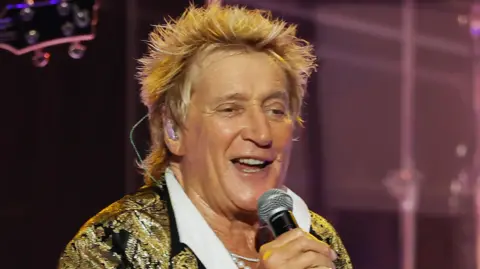 Sir Rod Stewart in a black and gold jacket, singing into a handheld microphone on stage on 31 July in Wantagh, New York