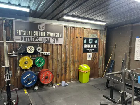 Charlotte Lillywhite/LDRS A corner of a gym with different colours and sizes of weights hanging on a wall bracket. There is other gym equipment visible either side of the weights. There is a green bin along the back wall and a sign that reads Physical Culture Gymnasium, Putney's local gym. 