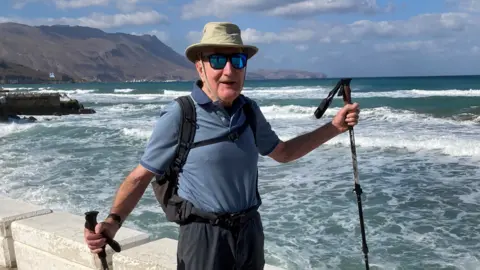Mike Patrick Mike Patrick is smiling with a scenic view in the background of the sea and mountains on a sunny day. He is wearing a blue polo shirt, sunglasses and a wide-brimmed hat, and is carrying walking poles.
